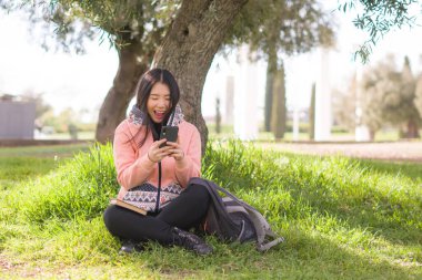 Şehir parkında cep telefonu kullanan mutlu bir Asyalı kadın. Yeşil çimlerin üzerinde oturan tatlı ve güzel bir Japon kız. Cep telefonuyla sosyal medyanın tadını çıkarıyor.