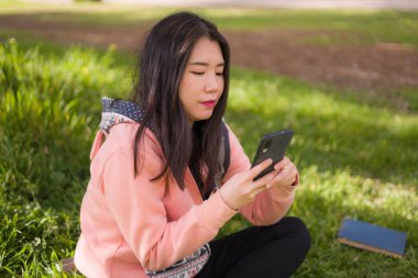 Şehir parkında cep telefonu kullanan mutlu bir Asyalı kadın. Yeşil çimlerin üzerinde oturan tatlı ve güzel bir Japon kız. Cep telefonuyla sosyal medyanın tadını çıkarıyor.