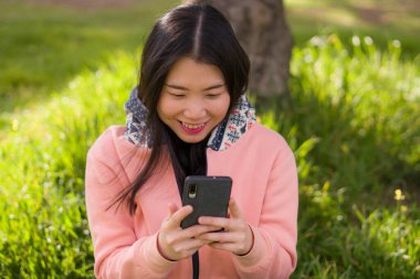 Şehir parkında cep telefonu kullanan mutlu Asyalı kadın. Yeşil çimlerin üzerinde oturan tatlı ve güzel Koreli kız. Cep telefonuyla sosyal medyanın keyfini çıkarıyor.