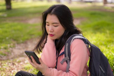 Şehir parkında cep telefonu kullanan mutlu Asyalı kadın. Yeşil çimlerin üzerinde oturan tatlı ve güzel Koreli kız. Cep telefonuyla sosyal medyanın keyfini çıkarıyor.