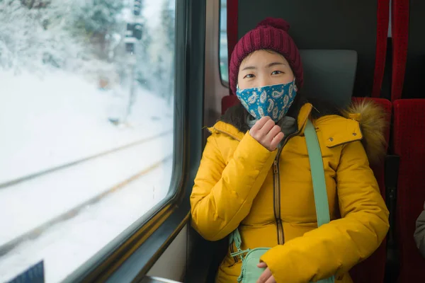 train traveling in times of covid19 - young happy and beautiful Asian Chinese woman in face mask going through snow landscape in winter holiday travel