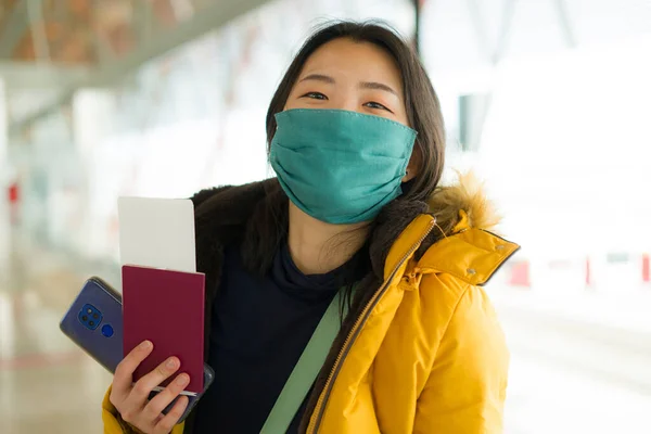Asian woman flying in covid19 times - lifestyle portrait of young happy and pretty Japanese girl in face mask and yellow jacket waiting on airport lounge ready for holiday trip