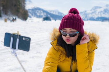 Kar altında kış tatili özçekimi - genç mutlu ve neşeli Asyalı Çinli kadın İsviçre Alplerinde soğuk karlı dağda cep telefonuyla kendi portresini çekiyor.