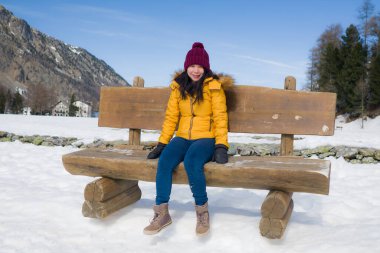 Sarı ceketli ve kışlık şapkalı genç mutlu ve güzel Asyalı kadın İsviçre Alp dağlarında karla kaplı donmuş gölün üzerindeki bankta neşeyle gülümsüyor.