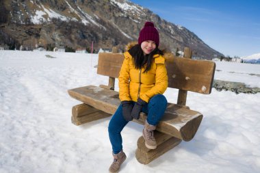 Sarı ceketli ve kışlık şapkalı genç mutlu ve güzel Asyalı kadın İsviçre Alp dağlarında karla kaplı donmuş gölün üzerindeki bankta neşeyle gülümsüyor.