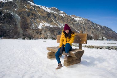Sarı ceketli ve kışlık şapkalı genç ve güzel Asyalı kadın İsviçre Alp dağlarında karla kaplı donmuş gölün üzerindeki bankta neşeyle gülümsüyor.