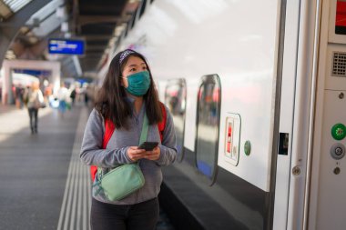 Asya 'lı sırt çantalı bir kız. 19 yaşında, mutlu ve güzel bir Japon kadın. Yüz maskesi ve sırt çantasıyla tren istasyonunda cep telefonunu kontrol ediyor. 