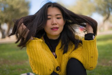 Sarı kazaklı, mutlu ve güzel Asyalı kadının hayat tarzı portresi bahar haftasonu yürüyüşünde ağacın yanındaki yeşil parkta eğleniyor.