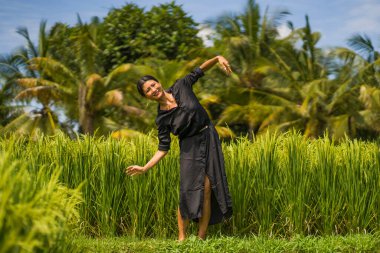 Çekici ve mutlu Asyalı bir kadının sanatsal portresi yeşil pirinç tarlasında zarif uzun elbiseler içinde tasasız bir şekilde dans ediyor. 