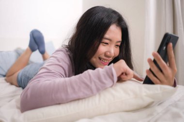 Cep telefonlu Asyalı bir kız. Yatak odasında neşeli gülümseyen internet uygulaması kullanarak genç mutlu ve çekici bir Çinli kadının portresi.