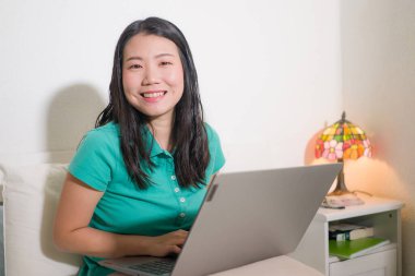 Yataktaki genç Asyalı kadın portresi - güzel ve mutlu Koreli kız rahat ve neşeli gülümseyen dizüstü bilgisayar kullanarak yatak odasında.