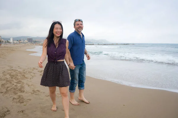 young beautiful and happy mixed ethnicity couple of Asian woman and Caucasian man relaxed and cheerful walking playful  on beach enjoying love and romantic holidays