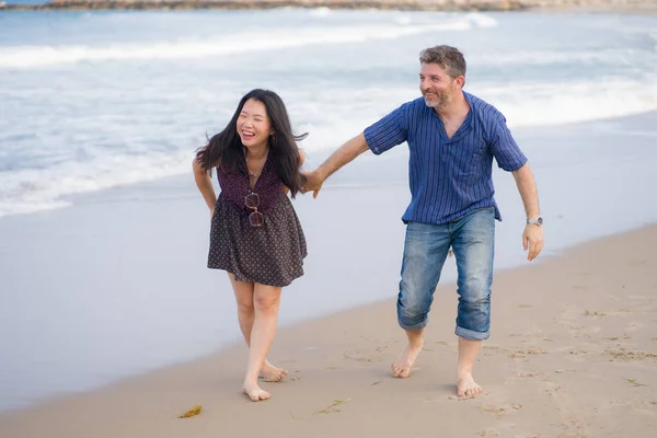 young beautiful and happy mixed ethnicity couple of Asian woman and Caucasian man relaxed and cheerful walking playful  on beach enjoying love and romantic holidays