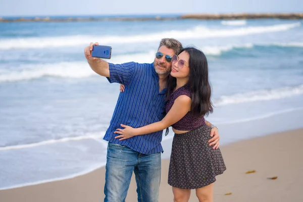 young happy and beautiful mixed ethnicity couple in love with Asian woman and Caucasian man taking sweet selfie with mobile phone on beach enjoying romantic holidays together
