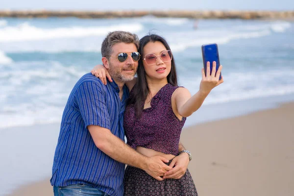 young happy and beautiful mixed ethnicity couple in love with Asian woman and Caucasian man taking sweet selfie with mobile phone on beach enjoying romantic holidays together