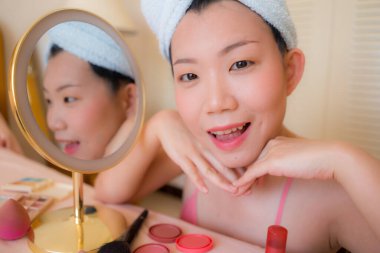 Güzel ve mutlu bir Asyalı kadın havluyla makyaj yapıyor aynaya bakıp gülümsüyor ve güzellik modasına hazırlanıyor.