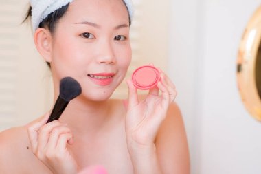 Güzel ve mutlu bir Asyalı kadın havluyla makyaj yapıyor aynaya bakıp gülümsüyor ve güzellik modasına hazırlanıyor.