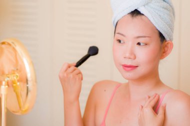 Güzel ve mutlu Asyalı kadın havluyla sarmalayıp makyaj yapıyor aynaya bakıp gülümsüyor ve güzellik modasına hazırlanıyor.