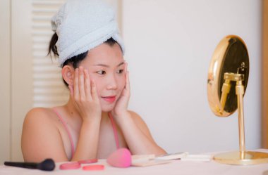 Güzel ve mutlu Asyalı kadın havluyla sarmalayıp makyaj yapıyor aynaya bakıp gülümsüyor ve güzellik modasına hazırlanıyor.