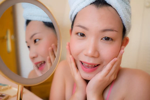 young beautiful and happy Asian Chinese woman with towel head wrap applying makeup looking to mirror smiling cheerful preparing for dating in beauty fashion concept