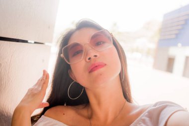 Güzel ve çekici Asyalı kadın cep telefonuyla selfie çekiyor. Güneş gözlüklü, güneşli bir günde sosyal medya ve internet paylaşımı için poz veriyor.