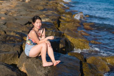 Deniz kenarındaki genç Asyalı kadının tatil tarzı portresi. Mutlu ve güzel Çinli kız plaj tatili yapıyor. Plaj kayalıklarında rahat ve neşeli.