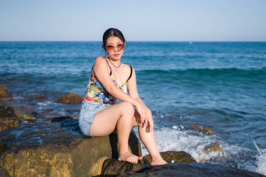 Deniz kenarındaki genç Asyalı kadının tatil tarzı portresi. Mutlu ve güzel Koreli kız plaj gezisinin keyfini çıkarıyor. Plaj kayalıklarında rahat ve neşeli.