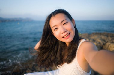 Genç mutlu ve güzel Asyalı kadın deniz kenarında cep telefonuyla selfie çekiyor. Japon kız yaz tatilinin tadını çıkarıyor. Plajda rahatça fotoğraf çekiyor.