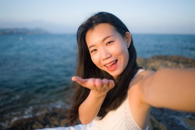 Genç ve güzel Asyalı kadın deniz kenarında cep telefonuyla selfie çekiyor. Koreli kız yaz tatilinin tadını çıkarıyor. Plajda rahatça fotoğraf çekiyor.