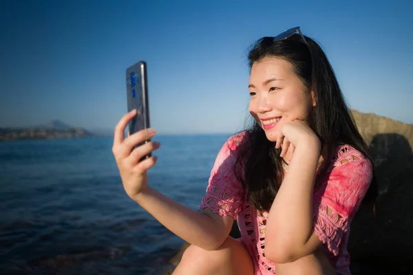행복하고 아름다운 아시아 소녀와 전화로 셀피를 복용하며 해변에서 편안하게 휴가를 — 스톡 사진