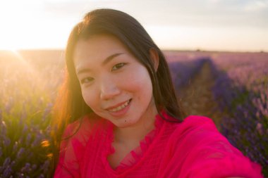 Genç, mutlu ve güzel bir Asyalı kadın yaz elbisesi giymiş. Mor lavanta çiçeklerinde cep telefonuyla günbatımı fotoğrafı çekiyor. Doğada ve romantik güzellik konseptinde.