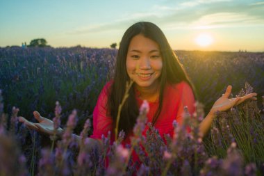 Genç ve güzel Asyalı yaz elbiseli kadın romantik güzellik ve özgürlük konseptiyle gün batımında mor lavanta çiçekleri tarlasında özgürce eğleniyor.