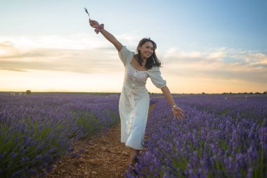 Açık havada, beyaz yaz elbiseli, mutlu ve çekici bir kadının romantik portresi seyahat ve tatil konseptindeki lavanta çiçekleri tarlasında tasasız eğleniyor.