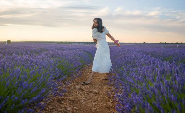 Açık havada, beyaz yaz elbiseli, mutlu ve çekici bir kadının romantik portresi seyahat ve tatil konseptindeki lavanta çiçekleri tarlasında tasasız eğleniyor.