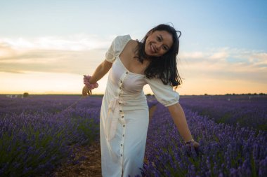 Açık havada, beyaz yaz elbiseli, mutlu ve çekici bir kadının romantik portresi seyahat ve tatil konseptindeki lavanta çiçekleri tarlasında tasasız eğleniyor.
