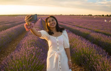 Açık hava romantik portresi beyaz yaz elbisesi içinde genç ve çekici bir kadın cep telefonuyla güzel lavanta çiçekleri tarlasında tatil konseptinde selfie çekiyor.