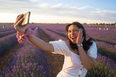 Açık hava romantik portresi beyaz yaz elbisesi içinde genç ve çekici bir kadın cep telefonuyla güzel lavanta çiçekleri tarlasında tatil konseptinde selfie çekiyor.