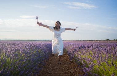 Açık havada, beyaz yaz elbiseli, mutlu ve çekici bir kadının romantik portresi seyahat ve tatil konseptindeki lavanta çiçekleri tarlasında tasasız eğleniyor.