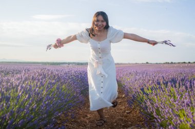 Açık havada, beyaz yaz elbiseli, mutlu ve çekici bir kadının romantik portresi seyahat ve tatil konseptindeki lavanta çiçekleri tarlasında tasasız eğleniyor.