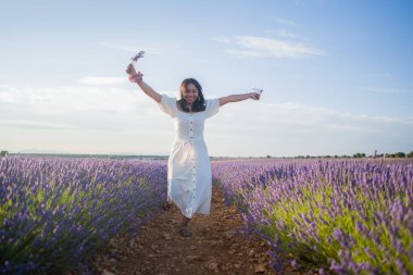 Açık havada, beyaz yaz elbiseli, mutlu ve çekici bir kadının romantik portresi seyahat ve tatil konseptindeki lavanta çiçekleri tarlasında tasasız eğleniyor.