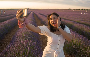 Açık hava romantik portresi beyaz yaz elbisesi içinde genç ve çekici bir kadın cep telefonuyla güzel lavanta çiçekleri tarlasında tatil konseptinde selfie çekiyor.