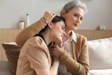 Orta yaşlı bir anne kızını kucaklıyor, hayal kırıklığına uğramış kızını gözleri kapalı, yanaklarına dokunuyor, evdeki kanepede oturuyor. Mutlu ve güvenilir ilişkiler. Aile kavramı