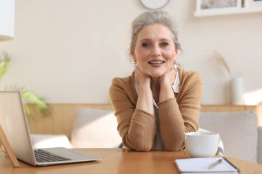 Middle aged woman sitting at a table with a laptop and looking at the camera smiling clipart