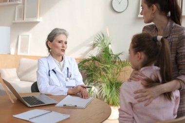 Anne ve çocuk doktor muayenehanesinde çocuk doktoruyla görüşüyorlar, hastanede masa başında oturuyorlar.