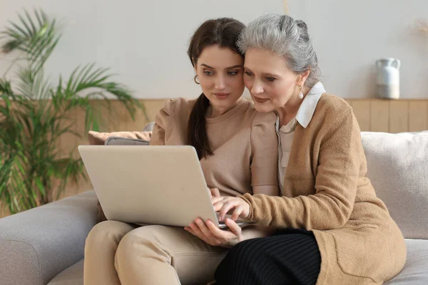 Happy elderly middle mother sitting on couch with her daughter, looking at laptop. Young woman showing video, photos to mommy, trusted relations. Family concept