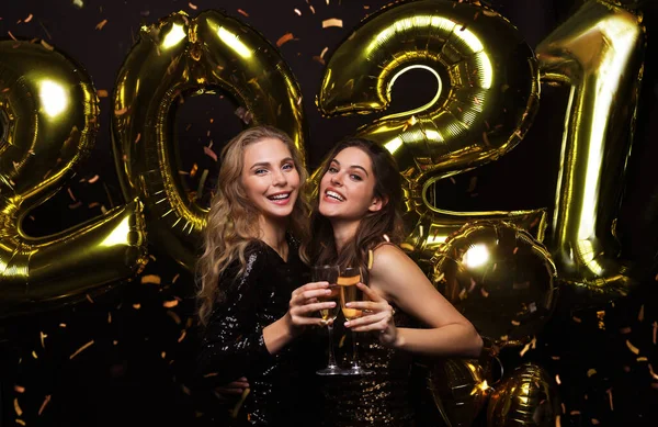 Две юные леди пьют шампанское. Изображение девушек с воздушными шарами на черном фоне, веселящихся на новогодней вечеринке 2021 года. — стоковое фото