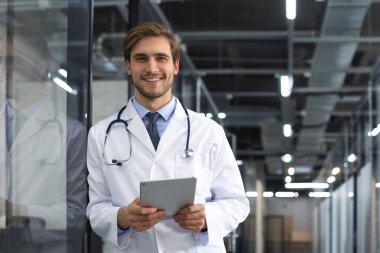 Hastane koridorunda yakışıklı genç doktor kameraya bakıyor, gülümsüyor.