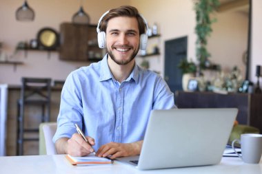 Kulaklıklı kendine güvenen işadamı evde dizüstü bilgisayarla otururken notlar veya mali raporlar yazıyor
