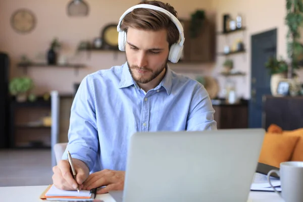 Confident businessman in earphones is writing notes or financial report while sitting at desk with laptop at home