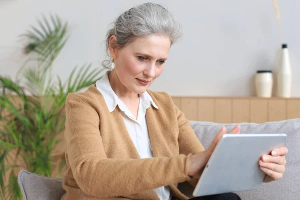 快乐的中年妇女坐在沙发上 使用电脑平板电脑应用程序 观看屏幕 在社交网络上阅读好消息 在网上购物或聊天 — 图库照片